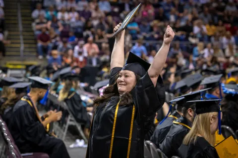 An excited UCCS graduate at Commencement holding her diploma cover in the air.